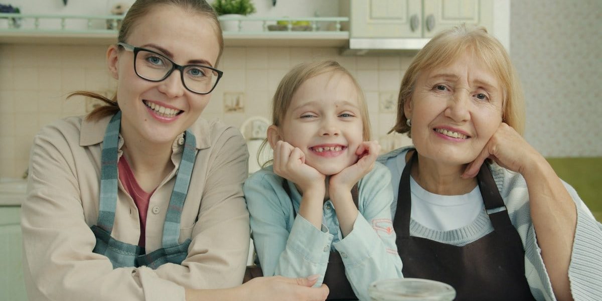 Portrait of joyful family grandmother, mother and daughter smiling in modern kitchen sitting together and looking at camera with happy faces.