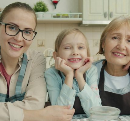 Portrait of joyful family grandmother, mother and daughter smiling in modern kitchen sitting together and looking at camera with happy faces.